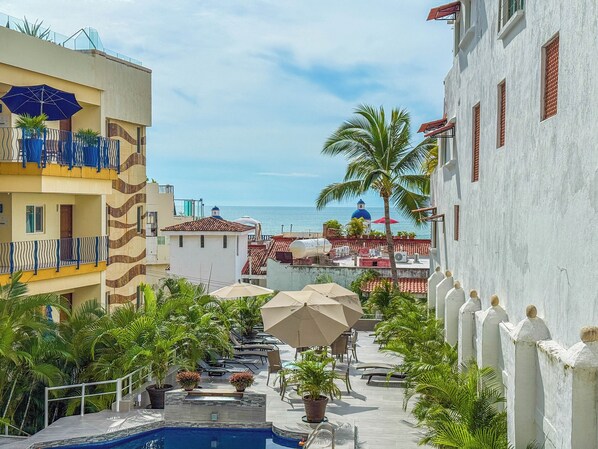 A pool area with lounge chairs and umbrellas, a view of the sea, and buildings with balconies and a dome structure. - Boana Torre Malibu #105 , Ocean view, romantic zone (Puerto Vallarta)