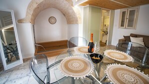 Un coin repas avec une table en verre, des chaises et une bouteille de champagne. On aperçoit en arrière-plan une arche en pierre et un canapé.