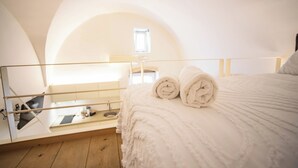 Une chambre avec un lit, deux serviettes enroulées, une petite fenêtre et un balcon avec une balustrade.