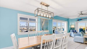 A dining area with a wooden table and white chairs, a ceiling fan, and a view of the sea through large windows. - The Bayside Oasis at Ocean Bliss | Boardwalk (Ocean City)