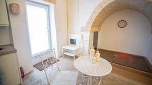 A room with a stone arch, a round table with bottles, a white chair, a small white table, and a door with a window. - Nivèra White Allure Maison · Suite Flùa – Ostuni (Ostuni)