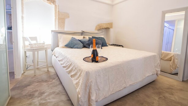 A bedroom with a bed, a bedside table, a chair, and a mirror. - Nivèra White Allure Maison · Suite Flùa – Ostuni (Ostuni)