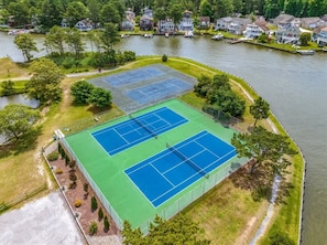 A tennis court complex with two courts, surrounded by a body of water and residential houses in the background. - Coastal Tides & Pines | Dog Friendly (Ocean Pines)