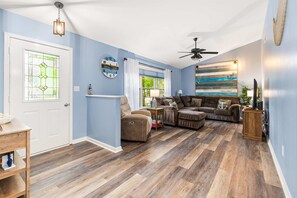A living room with a ceiling fan, a fireplace, a clock, and a large window with curtains. - Coastal Tides & Pines | Dog Friendly (Ocean Pines)