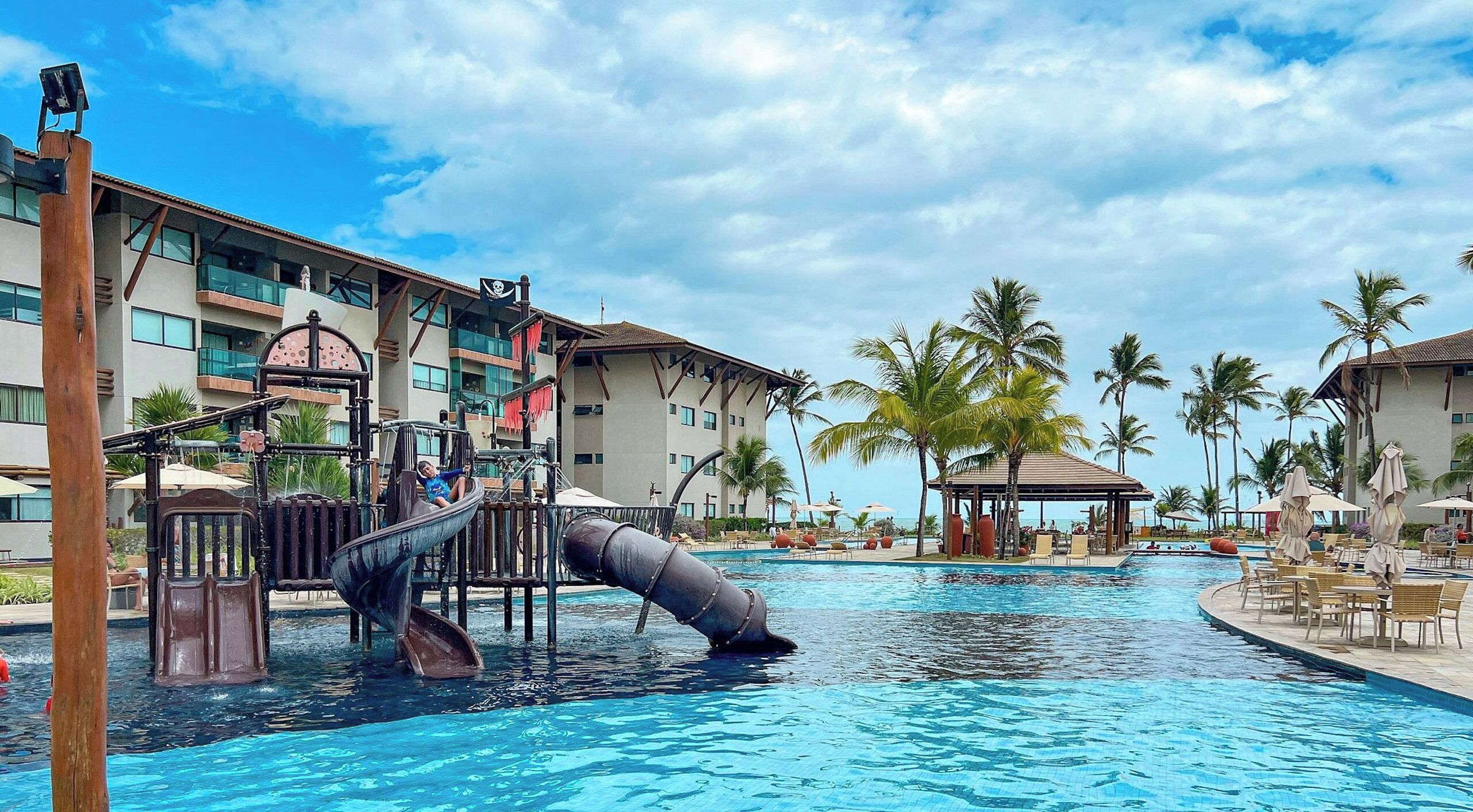 A swimming pool with a slide, palm trees, and a building with balconies.