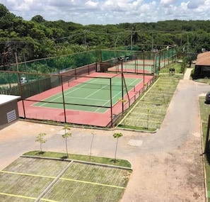 Una cancha de tenis con superficie verde y roja, rodeada por una valla, y un estacionamiento con algunos árboles.