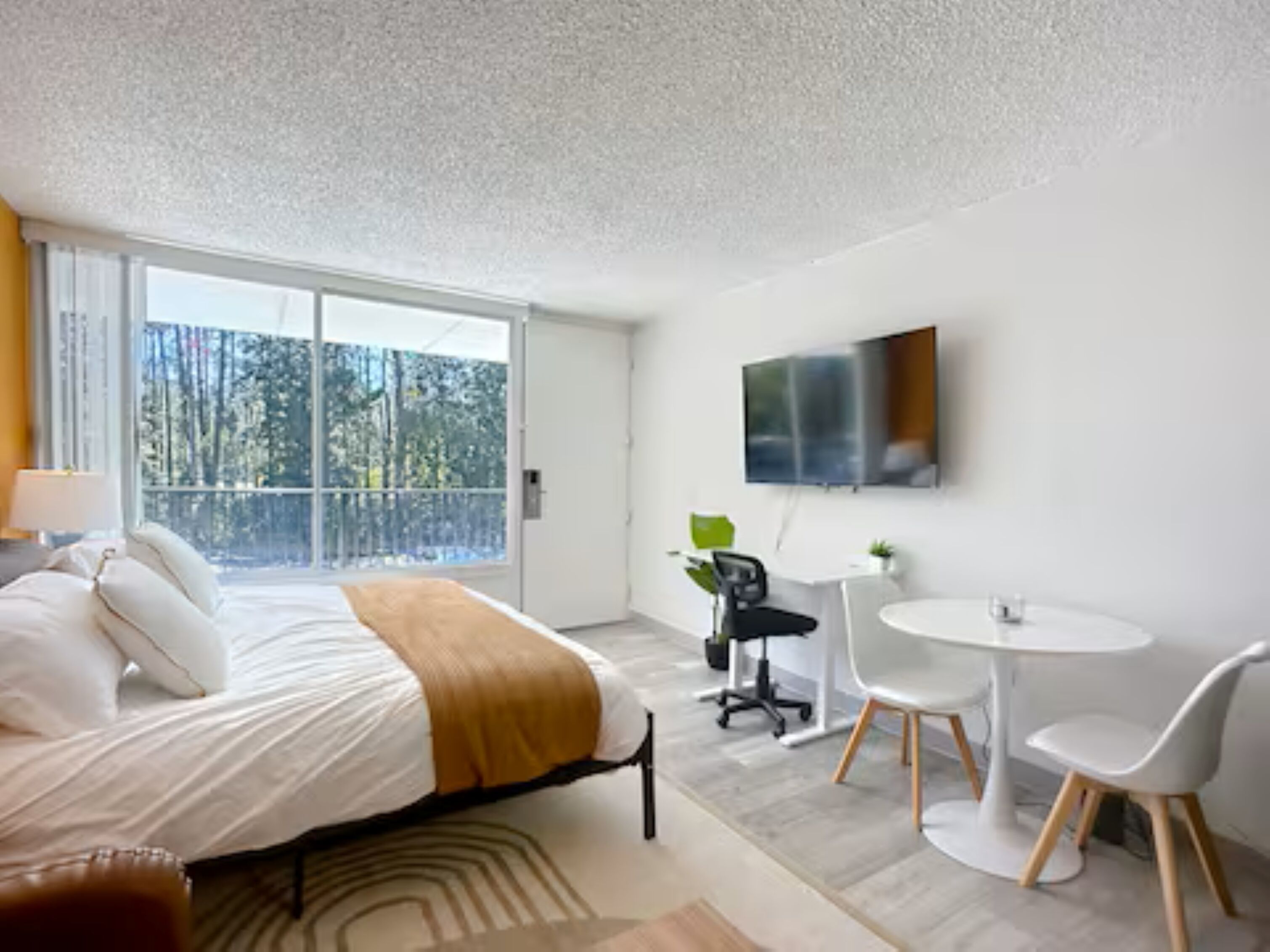 A modern hotel room with a bed, a desk, a chair, a TV, and a balcony with a view of trees.