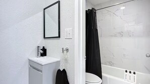 Un baño con lavamanos blanco, espejo y retrete. Hay una cortina de ducha negra y una bañera con mandos.