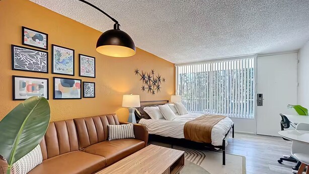 Un hotel moderno con un sofá de cuero marrón, una mesa de centro de madera, una cama con ropa blanca y un ventanal con persianas.