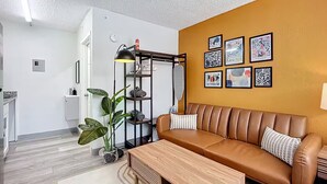 Una sala de estar moderna con un sofá de cuero marrón, una mesa de centro de madera y una estantería metálica negra con una lámpara. Hay cuadros enmarcados en la pared y una planta en el suelo.