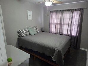 Ein Schlafzimmer mit einem Bett, einem Nachttisch, einer Lampe, einem Deckenventilator, einem Fenster mit Vorhängen und einer Wanddekoration.