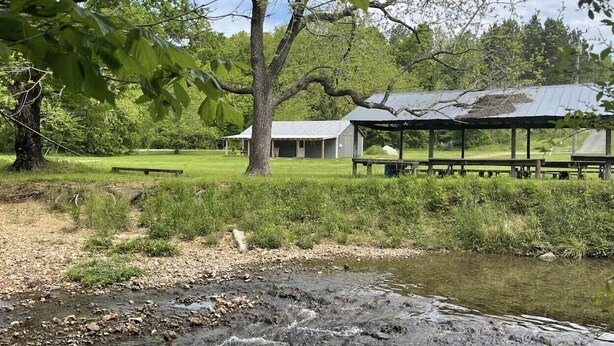 A rustic outdoor setting with a picnic area, a small stream, and a metal-roofed structure.