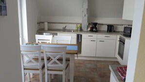 Een kleine keuken met witte kastjes, een eettafel met stoelen en een klein aanrecht met een koffiezetapparaat en een broodrooster.