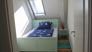 Een kleine kamer met een enkel bed, schuine wanden en een dakraam, een nachtkastje met lamp, en een kleurrijk gestreept vloerkleed.