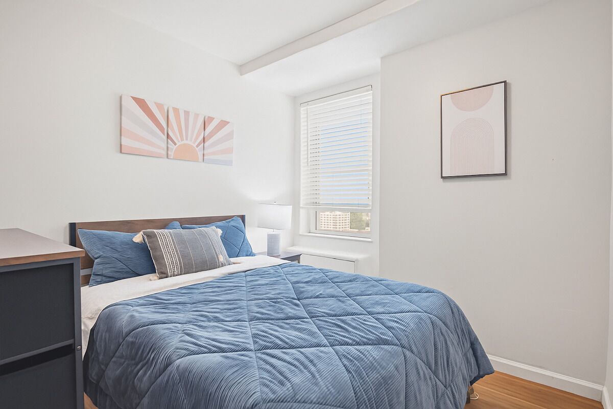 A bedroom with a bed, a nightstand, a window with blinds, and two wall art pieces.