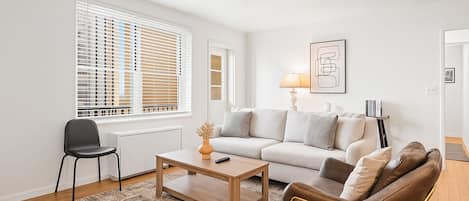 A modern living room with a white sofa, a brown leather chair, a wooden coffee table, and a black chair. There is a window with blinds, a lamp on the sofa, and a framed picture on the wall.