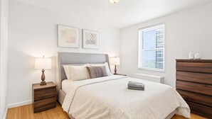 Ein Schlafzimmer mit einem Bett, zwei Nachttischen, einer Kommode, einem Fenster mit Jalousien und zwei Wandbildern.