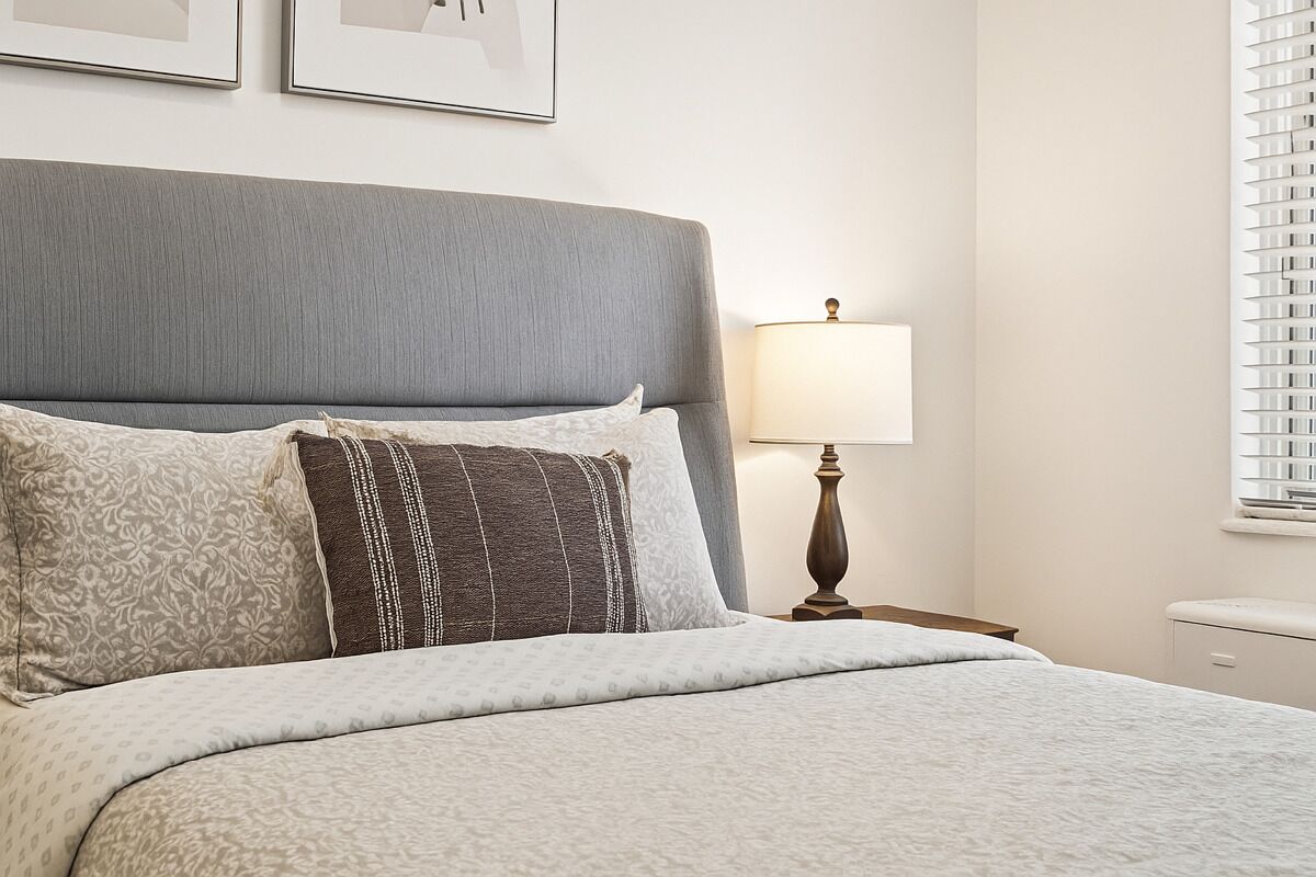 A neatly made bed with a grey headboard, a bedside lamp, and a window with blinds.