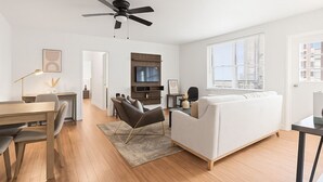 Ein modernes Wohnzimmer mit einer Couch, einem Essbereich mit Tisch und Stühlen, einem an der Wand montierten Fernseher und einem Deckenventilator.
