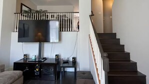 En moderne stue med et fladskærms-tv, et lavt møbel af mørkt træ og et tæppe med et mønster. En trappe med gelænder i træ fører til et andet rum.