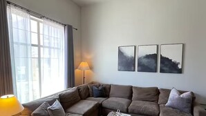 En stue med en hjørnesofa, et lille bord, en gulvlampe og en ventilator i loftet. Tre billeder hænger på væggen.