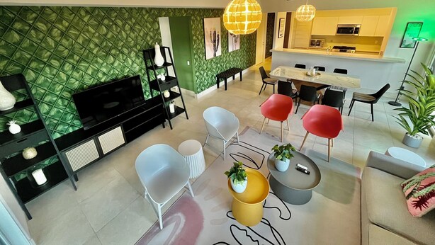 Een moderne woonkamer met een groene, gemêleerde wand, een zwarte entertainmentkast en een eethoek met een witte tafel en rode stoelen.