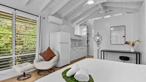 Ein modernes Schlafzimmer mit einem großen Bett, einem weißen Sessel, einer Küchenzeile und Blick auf Grün durch große Fenster.