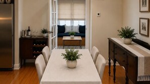 Een eetgedeelte met een tafel voor vier personen, een keuken met wijnflessen, en een zithoek met een bank en raam.