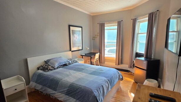 A bedroom with a bed, a wooden nightstand, a desk, a chair, a window with curtains, and a ceiling with a geometric pattern. - Kevinsrentals Dot Com (Providence)