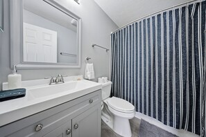 Ein Badezimmer mit weißem Waschbecken, Spiegel und Toilette. Ein gestreifter Duschvorhang und ein Handtuchhalter sind vorhanden.