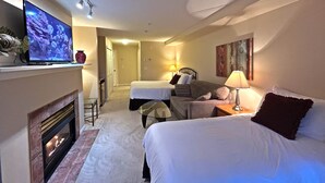 A hotel room with a bed, a sofa, a TV, a fireplace, and a hallway leading to another room. - Family Studio - 202B (Kelowna)