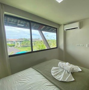 Een enkel bed met een wit laken en een handdoek, een raam met uitzicht op groen en gebouwen, en een airconditioningunit aan de muur.