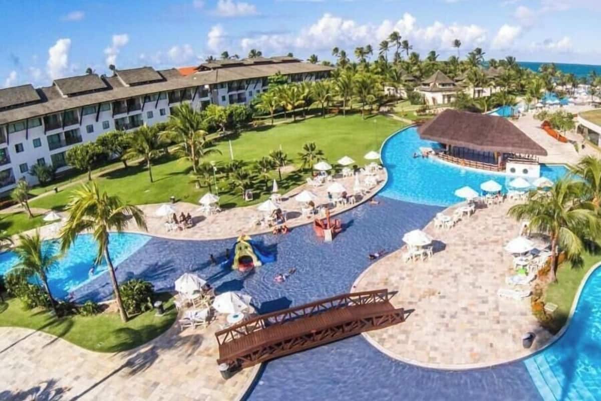 Un resort con una piscina grande, palmeras y un edificio con techo de paja.