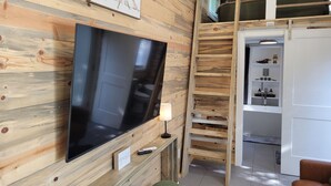 A room with a wooden wall, a flat-screen TV, a ladder, a green chair, and a white cabinet with shelves. - Tiny home in Van Alstyne with easy access to Hwy 75 and exceptional privacy. (Van Alstyne)