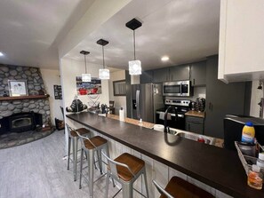A modern kitchen with a long island, bar stools, stainless steel appliances, pendant lights, and a stone fireplace. - Cal King, 2BR/2bth Hot Tub, Pool & Walk to town (Mammoth Lakes)