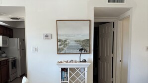 A clean, white interior with a kitchen area, a dining table with chairs, a white console table with books and decorative items, and a framed picture on the wall.