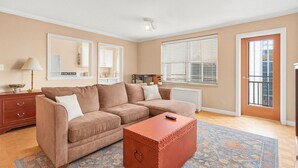 Ein Wohnzimmer mit braunem Sofa, Pouf in Rot, einem gemusterten Teppich, einer Lampe, einem Spiegel und einem Fenster mit Jalousien.