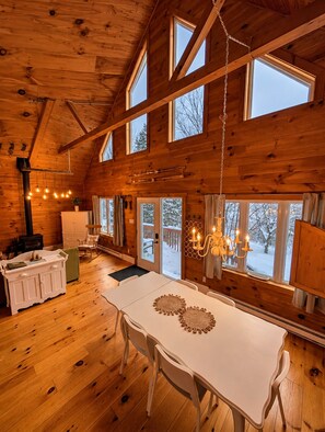A wooden cabin interior with a dining table, chairs, a chandelier, and large windows. - Full Guest Suite (Wakefield)