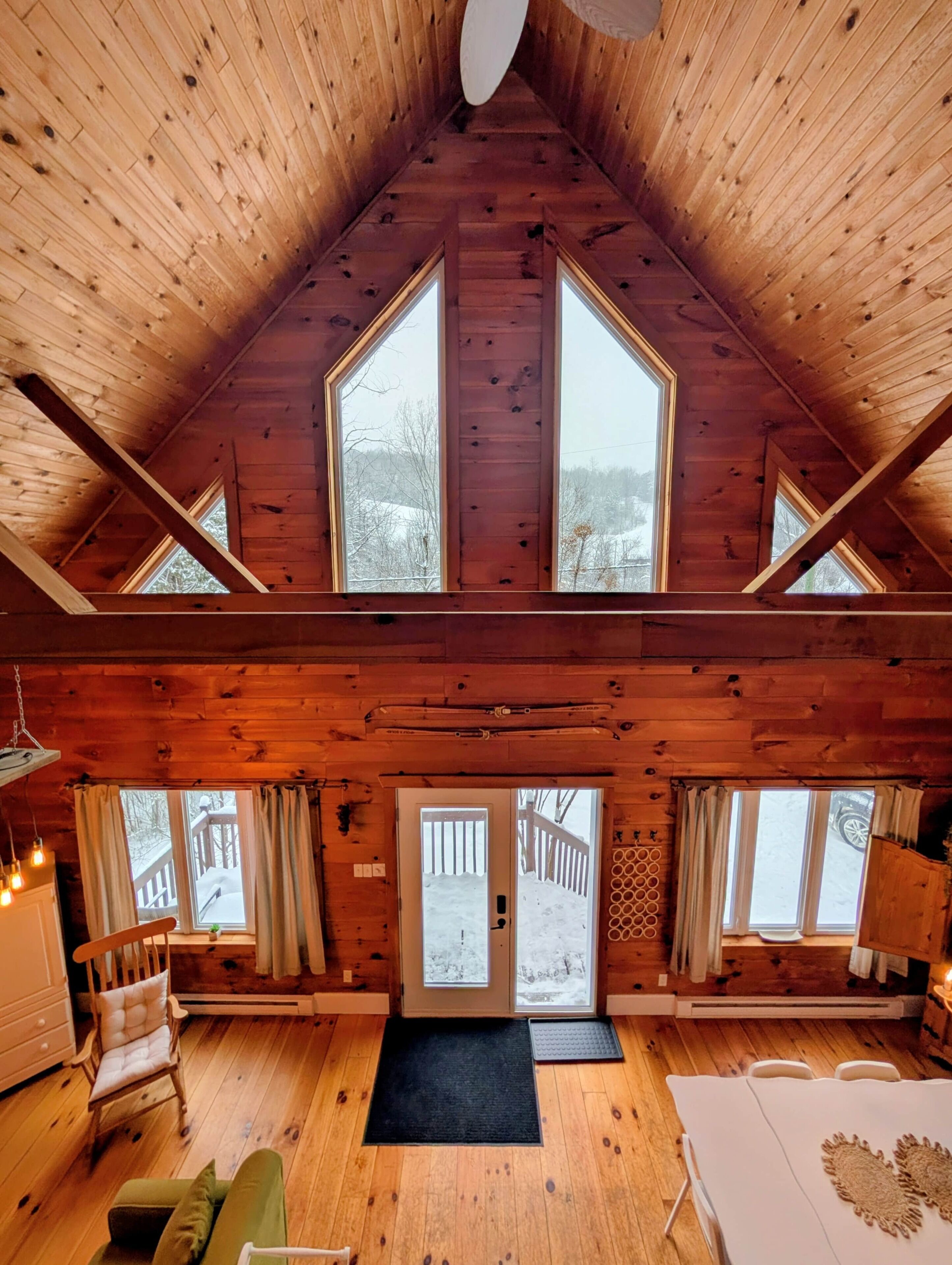 Une cabane en bois avec un plafond haut, de grandes fenêtres, un coin salon confortable et une vue sur une terrasse enneigée à l’extérieur.