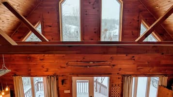 Une cabane en bois avec un plafond haut, de grandes fenêtres, un coin salon confortable et une vue sur une terrasse enneigée à l’extérieur.