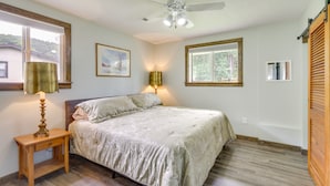 Un dormitorio con cama, una mesita de noche con lámpara, ventilador de techo y ventana con persianas.