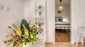 Ein Zimmer mit einem Bett, einem weißen Schreibtisch, einem weißen Stuhl, einer weißen Regalwand mit Pflanzen und einer Tür zu einem weiteren Raum.