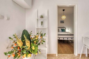Ein Zimmer mit einem Bett, einem weißen Schreibtisch, einem weißen Stuhl, einer weißen Regalwand mit Pflanzen und einer Tür zu einem weiteren Raum.