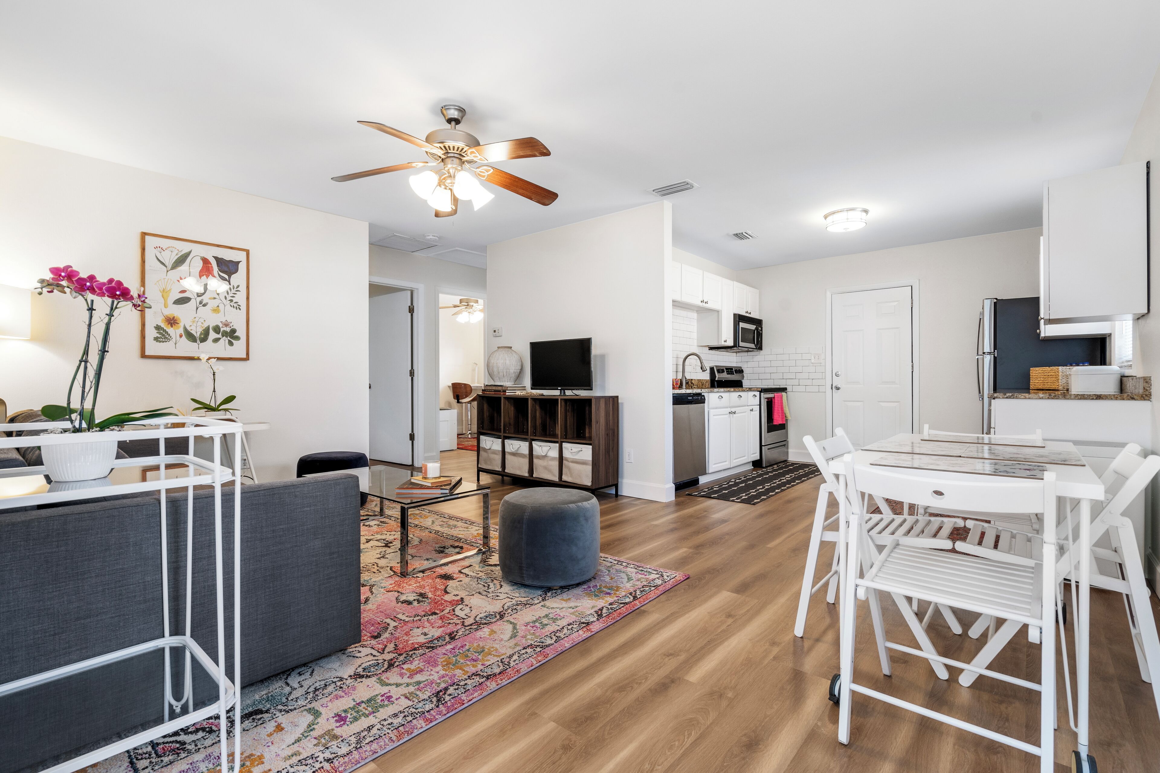 A modern living room with a gray sofa, a white dining table with chairs, a ceiling fan, and a kitchen area with white cabinets and a microwave.