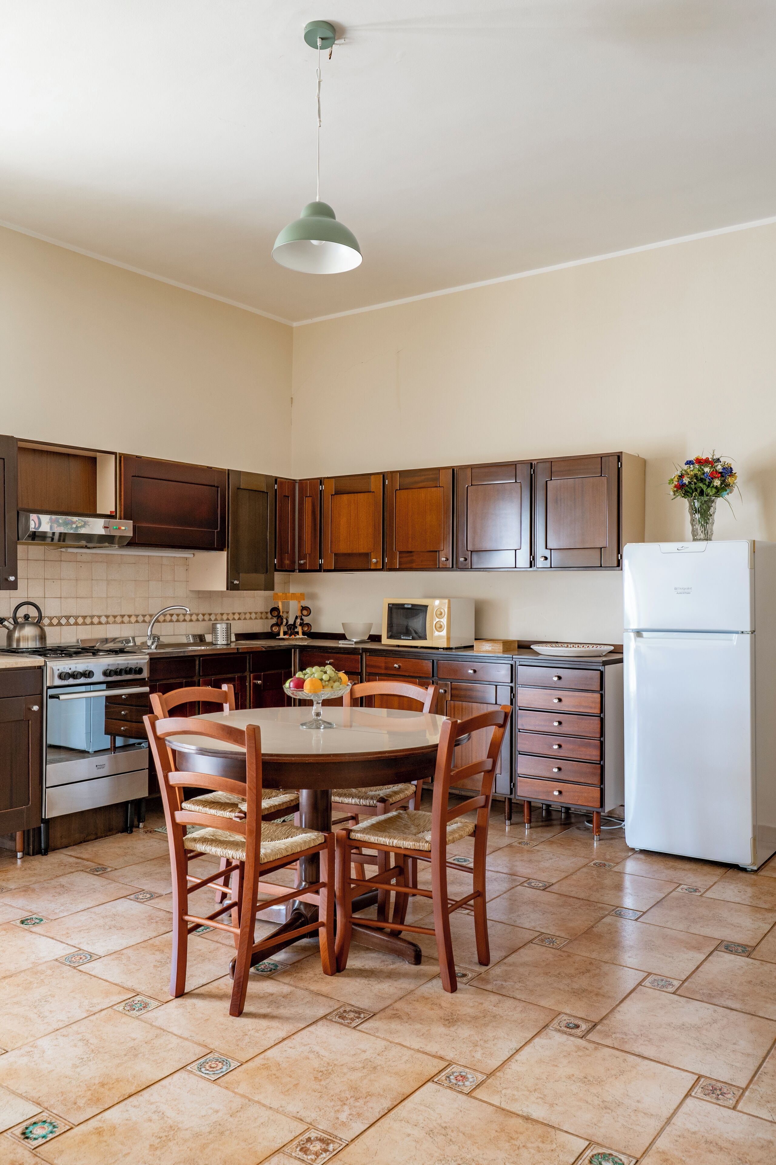 A kitchen with wooden cabinets, a dining table with chairs, a refrigerator, and a hanging light fixture.