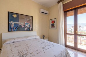 Une chambre d’hôtel avec un lit, un tableau, un appareil de climatisation, une table de chevet, une lampe et une vue sur la ville, visible par la porte d’un balcon.