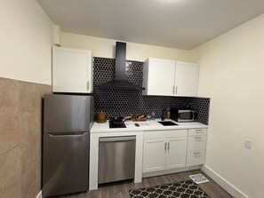 A compact kitchen with a stainless steel refrigerator, microwave, and dishwasher. It features white cabinets, a black hexagonal backsplash, and a small countertop with various kitchen items. - Modern farmhouse theme studio central Beamsville. Main Floor Suite. (Lincoln)