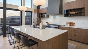 A modern kitchen with a large island, bar stools, and a view through large windows. - Whiskey Hill Luxury Retreat | Hot tub + Mountain Views (Vail)