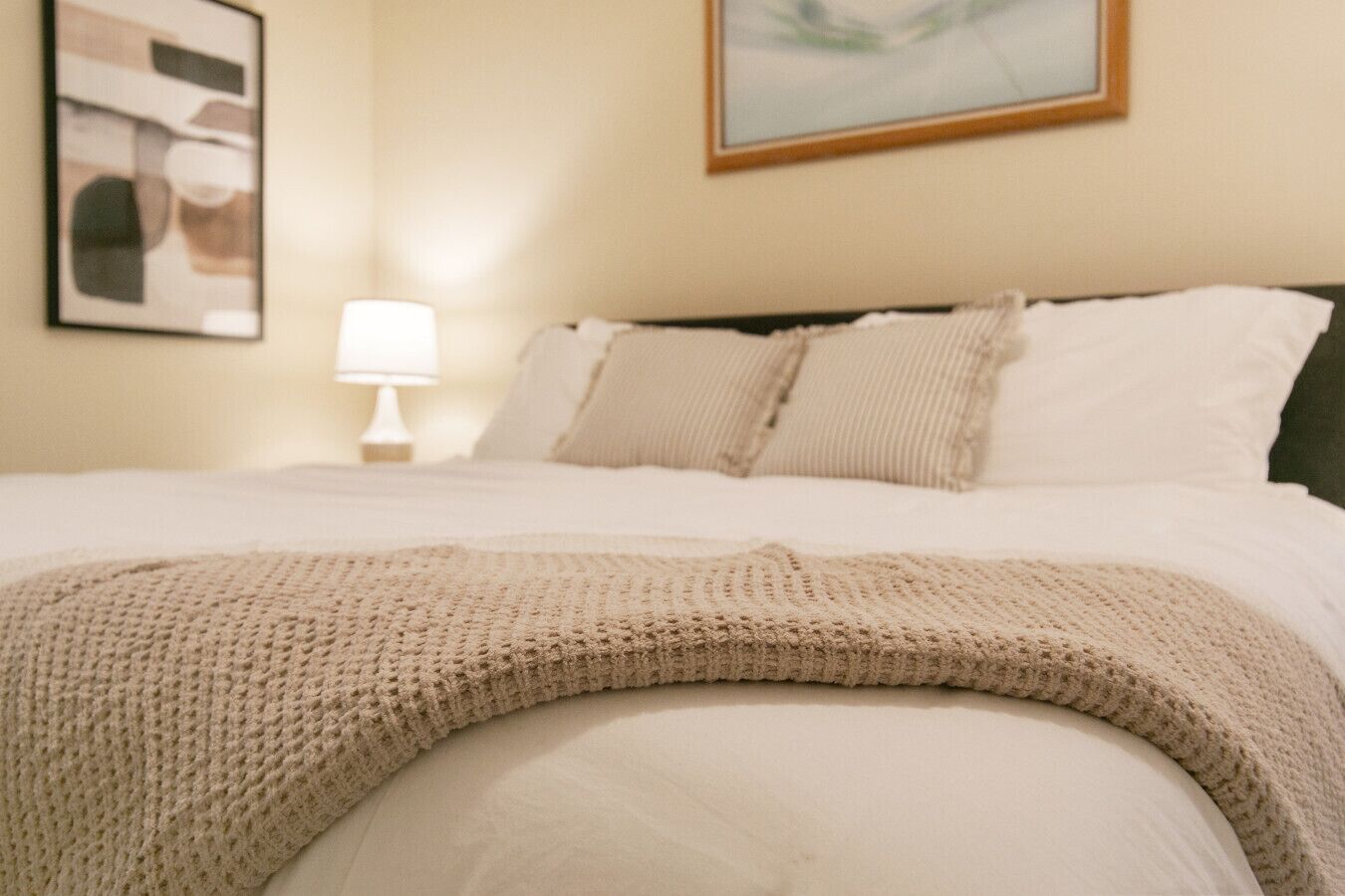 A neatly made bed with a knitted throw, a bedside lamp, and a framed abstract painting on the wall.
