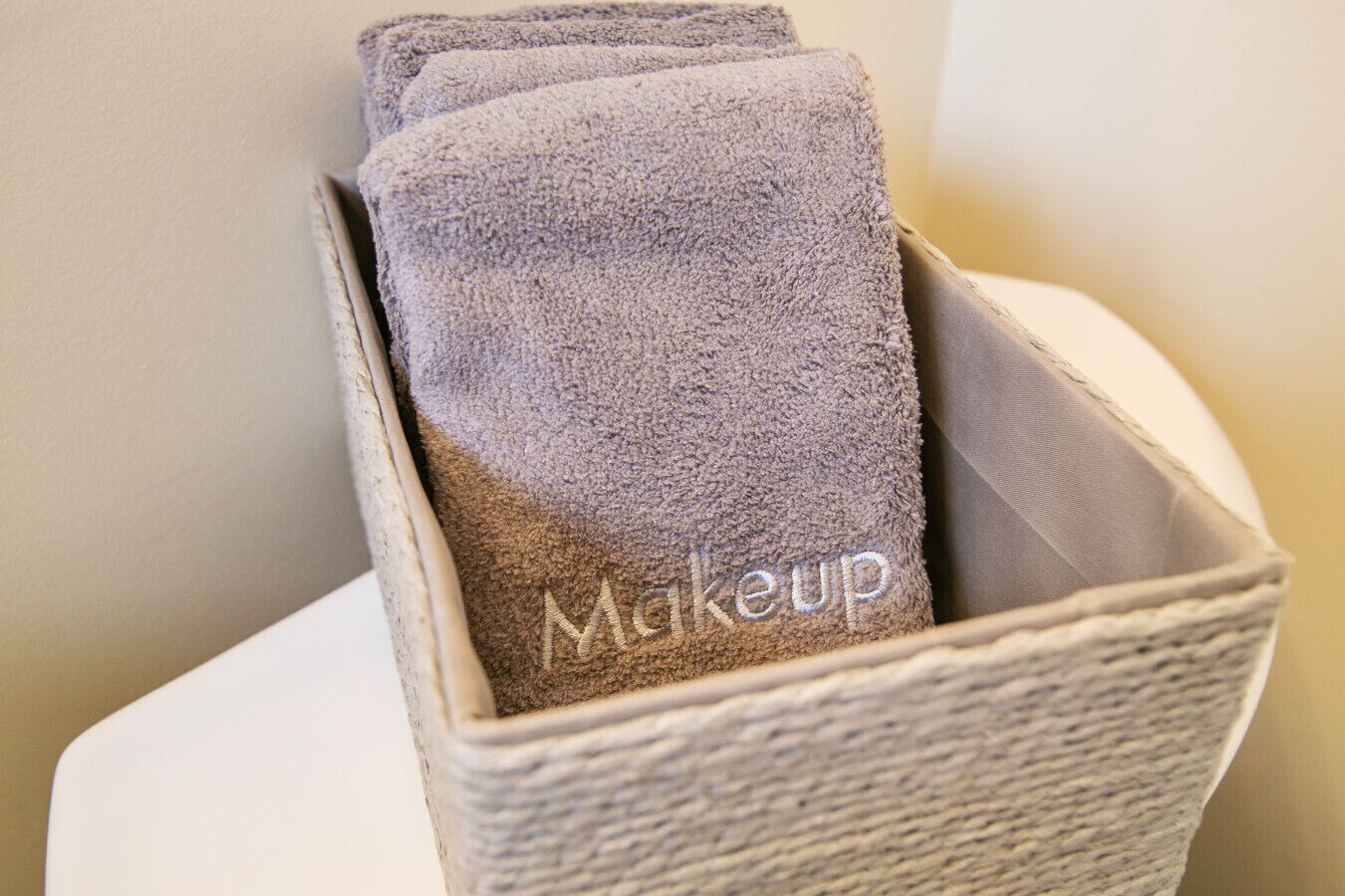 En opbevaringskasse med flettet overflade, der indeholder flere sammenfoldede håndklæder med broderet tekst 'makeup'.